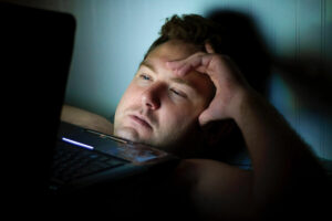 A man looking at a bright computer screen while lying in bed at night. Staring at device screens can keep you awake at night and affect your sleep patterns, causing anxiety.